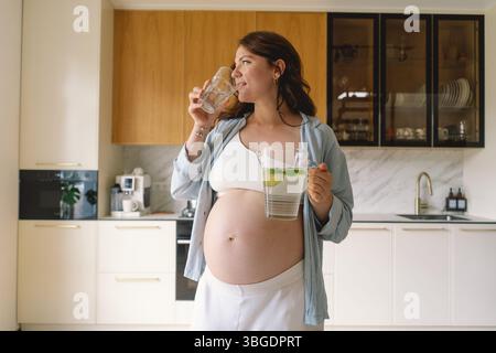 Schwangere Frau lächelt in der modernen Küche, während sie Krug und Glas Wasser mit Limettenscheiben hält Stockfoto