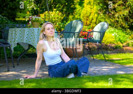 Eine blonde Frau in lässiger Kleidung sitzt auf dem Boden neben einer Gartenterrasse und genießt einen ruhigen Moment in der Sonne. Stockfoto
