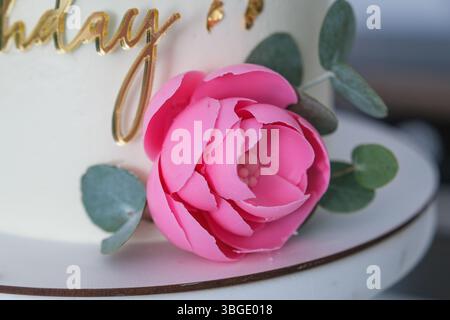 Eine rosa Blume steht auf einem weißen Kuchen mit den Worten „Happy Birthday“ in Gold Stockfoto