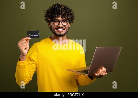 Junger Mann in lässiger gelber Kleidung, der Laptop und Kreditkarte vor khakifarbenem Hintergrund hält und Online-Shopping fördert Stockfoto