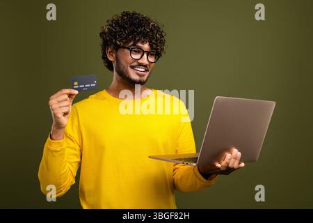 Junger fröhlicher Mann mit Kreditkarte und Laptop in stilvollem gelbem Outfit, Online-Shopping auf khakifarbenem Hintergrund Stockfoto