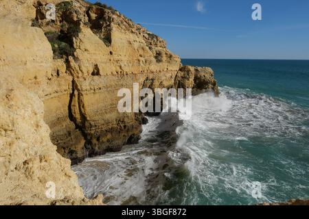 Wellen krachen an einer zerklüfteten Küste unter klarem blauen Himmel gegen goldene Klippen und fangen die Kraft und Schönheit der Natur ein. Stockfoto