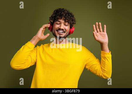 Fröhlicher junger Mann, der Musik über Kopfhörer hört, während er lächelt und gespendet, in einem leuchtend gelben Hemd und lässigem Stil Stockfoto