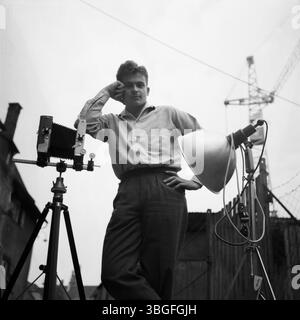 Der junge Fotograf Klaus Redenbacher lehnt lässig an seine Großformatkamera, neben ihm ein Studiolicht auf Stativ. Die Aufnahme entstand im Freien vor urbaner Kulisse mit Baukran – Deutschland, 1958. Stockfoto