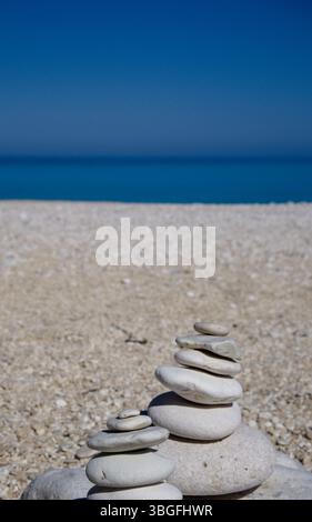 Weiße Kieselsteine stapeln sich an einem Sandstrand, blaues Meer im Hintergrund. Stockfoto