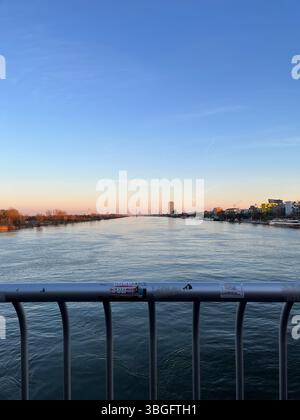 Blick von einer Brücke mit Blick auf einen ruhigen Fluss bei Sonnenuntergang mit Stadtblick und modernem Hochhaus in der Ferne. Stockfoto
