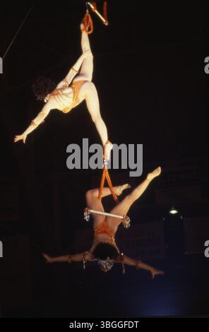 Foto von zwei weiblichen Trapezkünstlern beim 29. Jährlichen Aladdin Shrine Circus, der im Ohio State Fairgrounds Coliseum stattfindet. Ein Künstler hängt an einer horizontalen Leiste mit einem Fuß, während der andere an einer kurzen Leiste hängt, die am Fuß des ersten Künstlers befestigt ist. Stockfoto