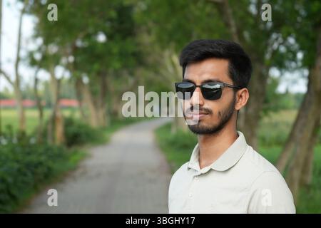 Junger Mann mit Sonnenbrille, der neben einem Baum auf einem grünen Land steht Stockfoto