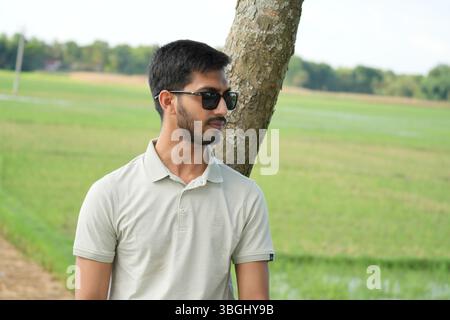 Junger Mann mit Sonnenbrille, der neben einem Baum auf einem grünen Land steht Stockfoto