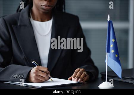 Abgeschnittene Aufnahme einer jungen erwachsenen schwarzen Politikerin, die mit Stift auf Papier schreibt, während sie am Schreibtisch neben der kleinen Flagge der Europäischen Union im politischen Amt sitzt Stockfoto