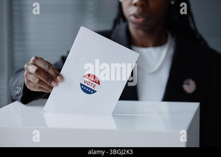 Zugeschnittene Aufnahme einer schwarzen Wahlteilnehmerin im Business-Anzug, die Stimmzettel in die Wahlbox wirft und einen Umschlag mit Stimmaufkleber in der Wahlstation hält Stockfoto