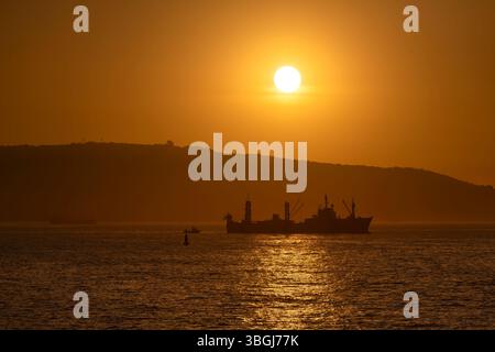 Ein Fischerboot fährt durch die ruhigen Gewässer von Valparaiso, Chile, während die Sonne hinter einem fernen Hügel untergeht und einen warmen goldenen Glanz über den Hafen wirft Stockfoto