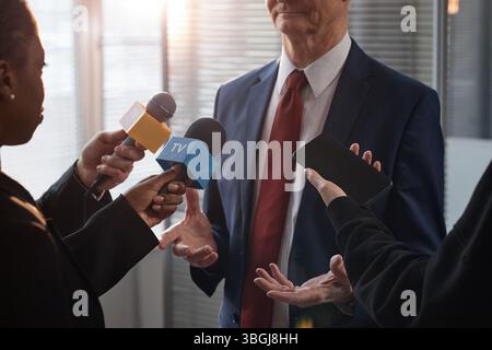 Abgeschnittene Aufnahme eines männlichen Politikers mittleren Alters, der mit Händen Gesten macht, während er von einer Gruppe von Journalisten, die Mikrofone und Smartphone im Büro halten, interviewt wird Stockfoto