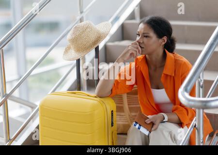 Nachdenkliche Schwarze Frau, Die Mit Gepäck Auf Der Treppe Im Flughafen Sitzt Stockfoto