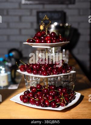 Dreistufiger, weiß-goldener Serviertisch gefüllt mit frischen roten Kirschen, angeordnet auf einer Holzoberfläche in gemütlicher Umgebung. Stockfoto