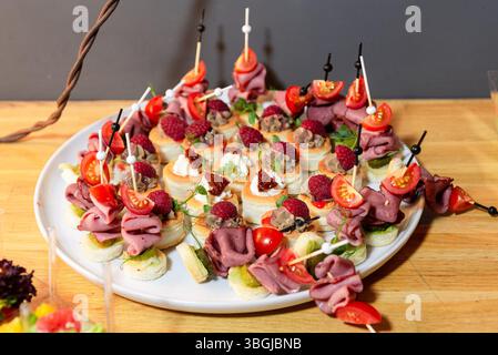 Elegante Platte mit verschiedenen Gourmet-Kanapees, belegt mit Aufschnitt, frischen Himbeeren, Kirschtomaten und Creme auf einem Holztisch. Stockfoto