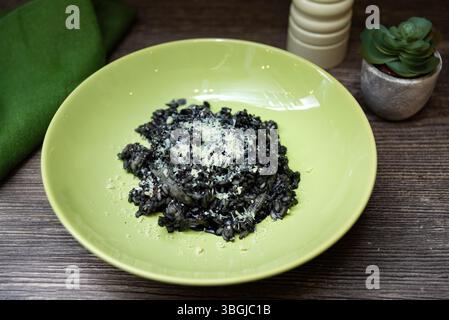 Nahaufnahme mit Tintenfisch-Tintenrisotto mit geriebenem Parmesan, serviert in einer grünen Schüssel auf einem rustikalen Holztisch mit Dekoelementen. Stockfoto