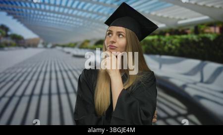 Eine junge Frau in Abschlusskleidung steht nachdenklich in einer Stadtstraße unter moderner Architektur und denkt über ihre Zukunft nach. Stockfoto