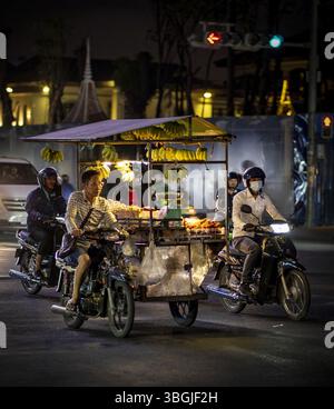 Eine geschäftige Nachtszene mit einem Straßenverkäufer, der Bananen aus einem Wagen verkauft, umgeben von Motorrädern und Stadtlichtern. Die Atmosphäre ist lebhaft und man Stockfoto