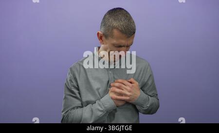 Kaukasischer Mann in grauem Hemd hält Brust mit emotionalem Ausdruck vor einem lila isolierten Hintergrund. Stockfoto
