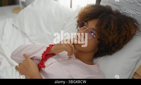 Junge afroamerikanische Frau mit lockigem Haar liegt nachdenklich auf dem Bett im gemütlichen Schlafzimmer, trägt eine Brille und verkörpert eine entspannte Inneneinrichtung. Stockfoto