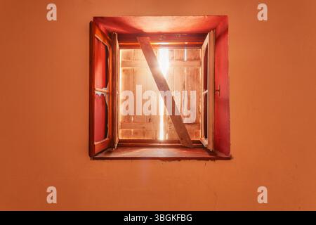Altes rotes Fenster mit Rollenverschluss auf verlassenem alpinen Gebäude, mit gefiltertem Licht durch Holzrisse Stockfoto
