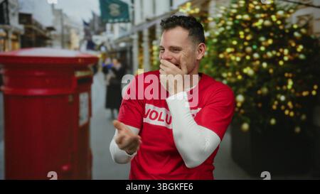 Mann lacht fröhlich in der Stadtstraße, trägt ein rotes Hemd mit Weihnachtsdekoration und einen britischen Briefkasten im Hintergrund während der Weihnachtszeit. Stockfoto