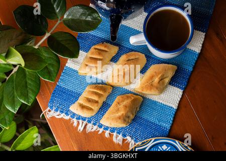 Eine traditionelle paraguayische Frühstücksszene, aufgenommen von oben, mit sechs Chipas auf einem gewebten blau-weißen Tischset, einer dampfenden Tasse Cocido in einem Stockfoto