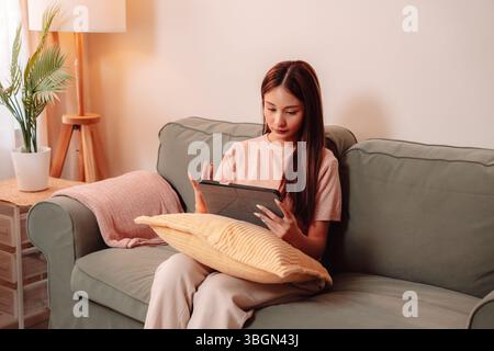 Frau sitzend Sofa Couch Medientechnologie Tablet Lesen Wohnzimmer, asiatische Frau genießt Online-Inhalte und entspannt zu Hause in Komfort, verbunden Stockfoto