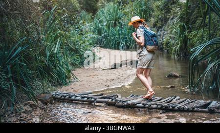 Junge Frau in beigefarbenem Hemd, Shorts und Strohhaw, Rucksack auf den Schultern, Wandern im Regenwald-Dschungel, Spaziergang über eine niedrige Holzbrücke am kleinen Bach in der Nähe Stockfoto