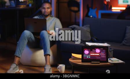 Black IT Professional in Loft arbeitet an Cybersicherheit mit Gadgets, geht Hacking-Versuchen, Verschlüsselung und Cyberabwehr an. Der Bildschirm zeigt Warnungen zu Sicherheitsverletzungen und Datensicherheitsrisiken an. Kamera B.. Stockfoto