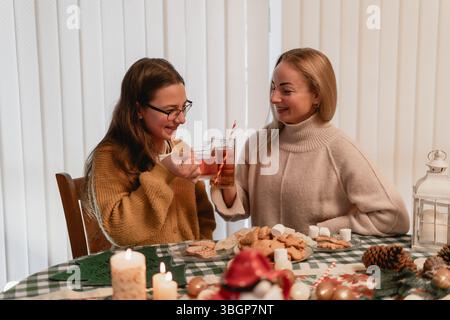In einem warmen Feiertag mit Tee toasten zwei Frauen mit Lachliebe und süßen Keksen, umgeben von Marshmallows bei Kerzenschein und festlicher Ruhe Stockfoto
