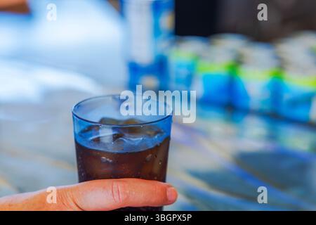 Handhaltendes Kunststoffglas, gefüllt mit Eiskola vor farbenfrohem Hintergrund mit verschwommenen Flaschen. Aruba. Stockfoto