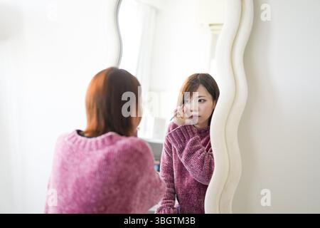 In einem sanft beleuchteten, sauberen Raum steht eine schwangere Japanerin Anfang 20 mit kurzen braunen Haaren neben einem Spiegel und trägt beim Tragen einen Lidstrich auf Stockfoto
