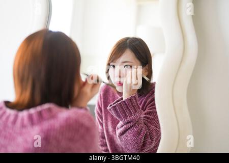 In einem sanft beleuchteten, sauberen Raum steht eine schwangere Japanerin Anfang 20 mit kurzen braunen Haaren neben einem Spiegel und trägt beim Tragen einen Lidstrich auf Stockfoto