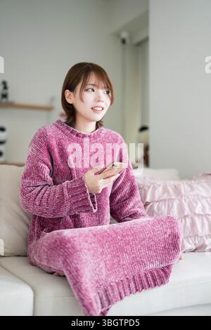 In einem sauberen, einfachen Raum mit sanftem Licht trägt eine schwangere Japanerin Anfang 20 mit kurzen braunen Haaren ein rosafarbenes Strickkleid. Sie sitzt auf einem Stockfoto