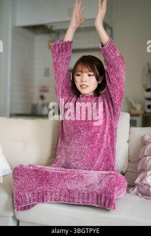 In einem ruhigen, gut beleuchteten Raum sitzt eine schwangere Japanerin Anfang 20 mit kurzen braunen Haaren in einem rosa Strickkleid auf einem Sofa und übt Stockfoto