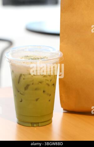 Eine Nahaufnahme eines erfrischenden, eiskalten Matcha-Latte in einer klaren Tasse mit Strohhalm, der neben einer braunen Papiertüte auf einem Holztisch in einer hellen Café-Atmosphäre sitzt Stockfoto