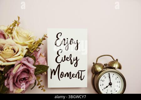 Genießen Sie jeden Moment Text auf Papierkarte mit Wecker auf rosa Hintergrund Stockfoto