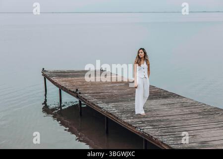 Asiatische Frau in weißem Outfit steht auf hölzernem Pier, ruhiger See, friedlich bewölkt, von hinten betrachtet mit kontemplativer Stimmung, Achtsamkeit, Reisen Stockfoto