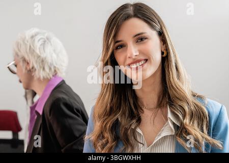 Porträt einer glücklichen jungen Geschäftsfrau, die in die Kamera lächelt. Ein leitender Kollege ist im Hintergrund verschwommen und steht für eine moderne, mu Stockfoto