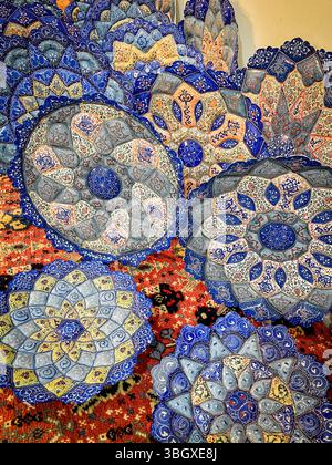 Aufwendig gemusterte persische Keramikplatten auf einem lebendigen Teppich mit handbemalten Blumenmotiven in satten Blau-, Orange- und Grautönen Stockfoto