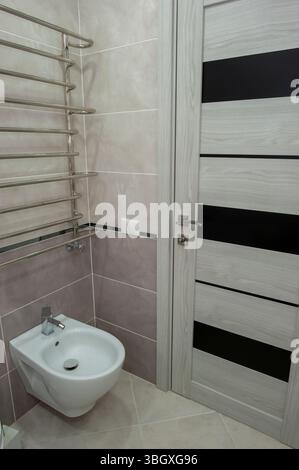 Großes, helles, neues Badezimmer in hellen Farben mit grauem, warmem Fliesenboden und weißer Tür gegen das unvollendete Zimmer. Stockfoto