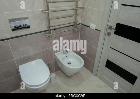 Großes, helles, neues Badezimmer in hellen Farben mit grauem, warmem Fliesenboden und weißer Tür gegen das unvollendete Zimmer. Stockfoto