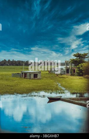 Eine ruhige Landschaft mit zwei bescheidenen Häusern, üppigen grünen Feldern, die teilweise in Wasser getaucht sind, und einem kleinen Boot, das auf der ruhigen Reflektion ruht Stockfoto
