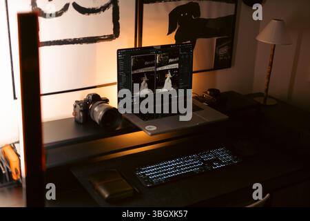 Der schwache Schreibtisch eines Fotografen mit einem Laptop, auf dem die bearbeiteten Hochzeitsfotos angezeigt werden, eine Digitalkamera mit Objektiv, eine externe Tastatur, ein Stockfoto