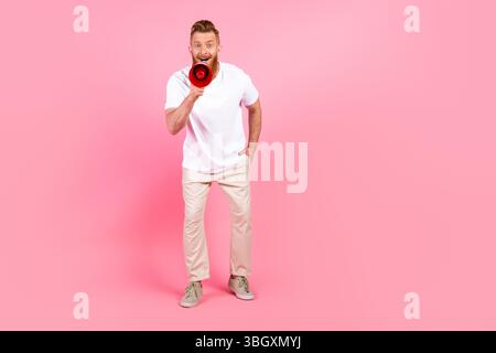 Junger Mann mit Bart und weißem T-Shirt, der fröhlich durch ein Megaphon vor rosafarbenem Hintergrund brüllt, fröhlich und stilvoll Stockfoto