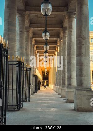 Eine lange, sonnendurchflutete Säulenhalle mit sich wiederholenden klassischen Säulen und hängenden Laternen schafft ein starkes Gefühl der Perspektive. Eine Einzelfigur läuft in der Dista Stockfoto