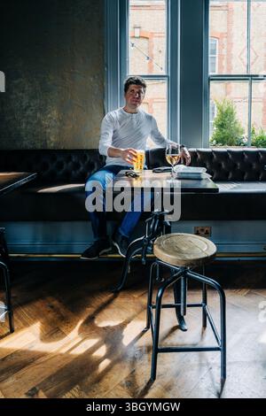 Mann sitzt am Fenster mit Bier und Wein auf einem Holztisch Stockfoto
