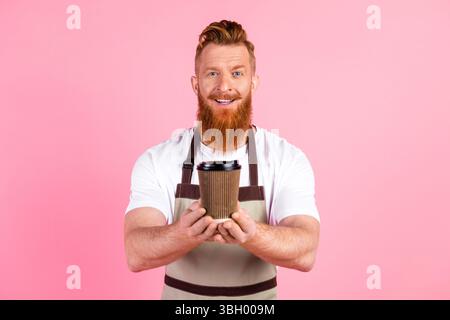 Lächelnder junger Mann mit rotem Bart, der eine Kaffeetasse in einer trendsetzenden Schürze vor einem leuchtend rosa Hintergrund hält Stockfoto